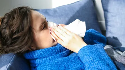 Žena v modrém svetru leží na gauči a smrkáním do kapesníku se snaží ulevit od rýmy